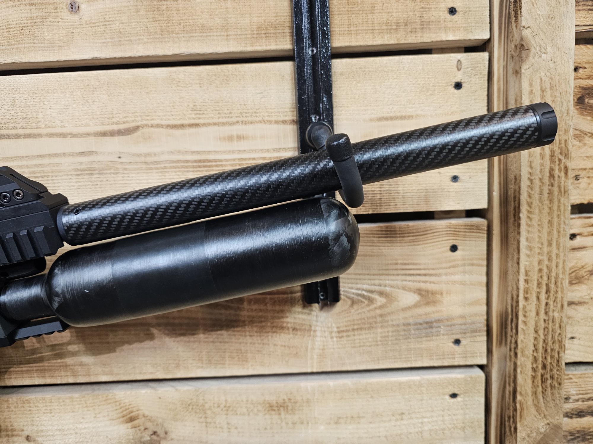 Air gun featuring a carbon fiber barrel and a black air cylinder mounted on a wooden wall.