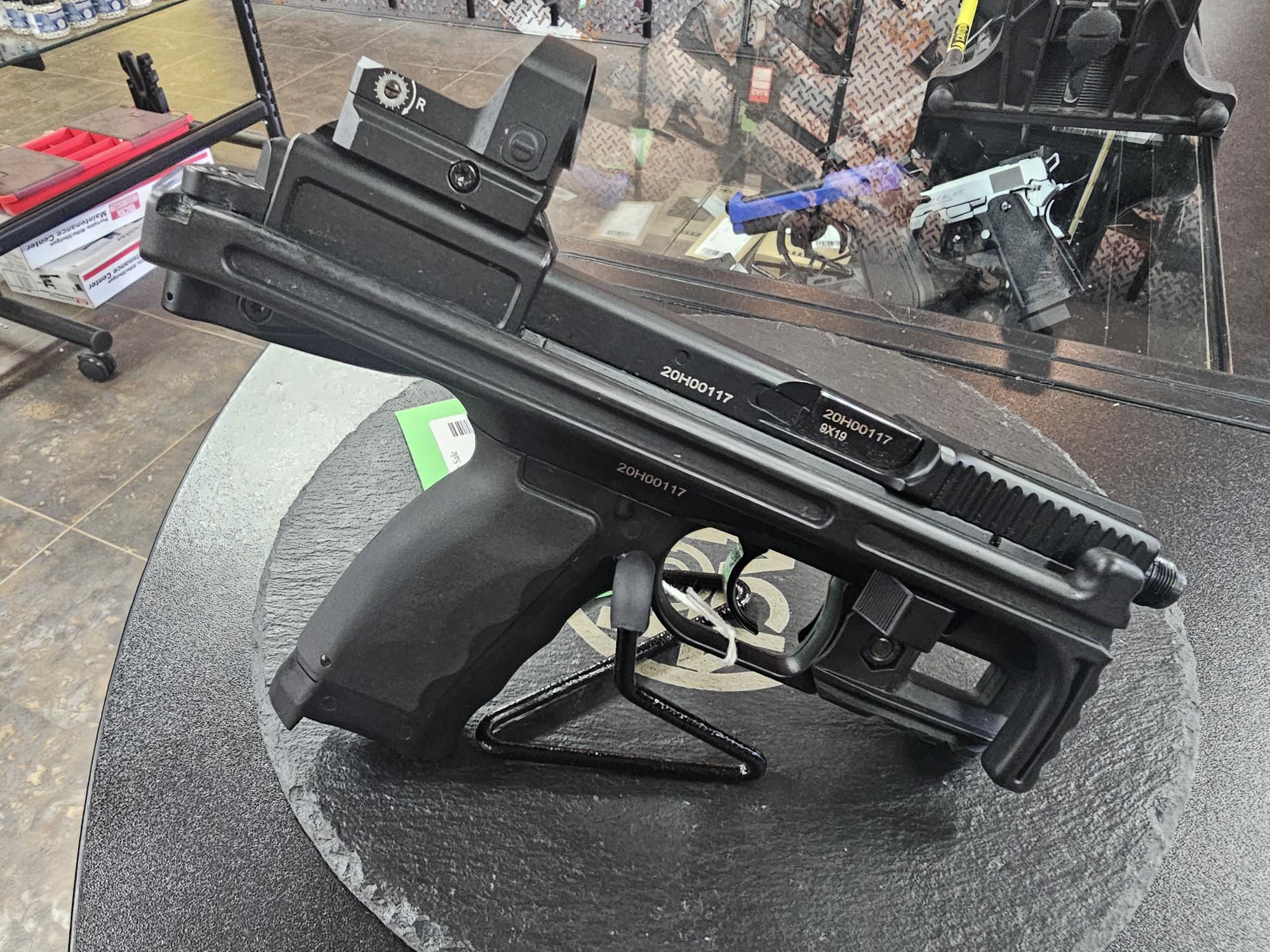 Displayed B&T USW airsoft pistol with a mounted red-dot sight, black finish and visible compact frame on a stand