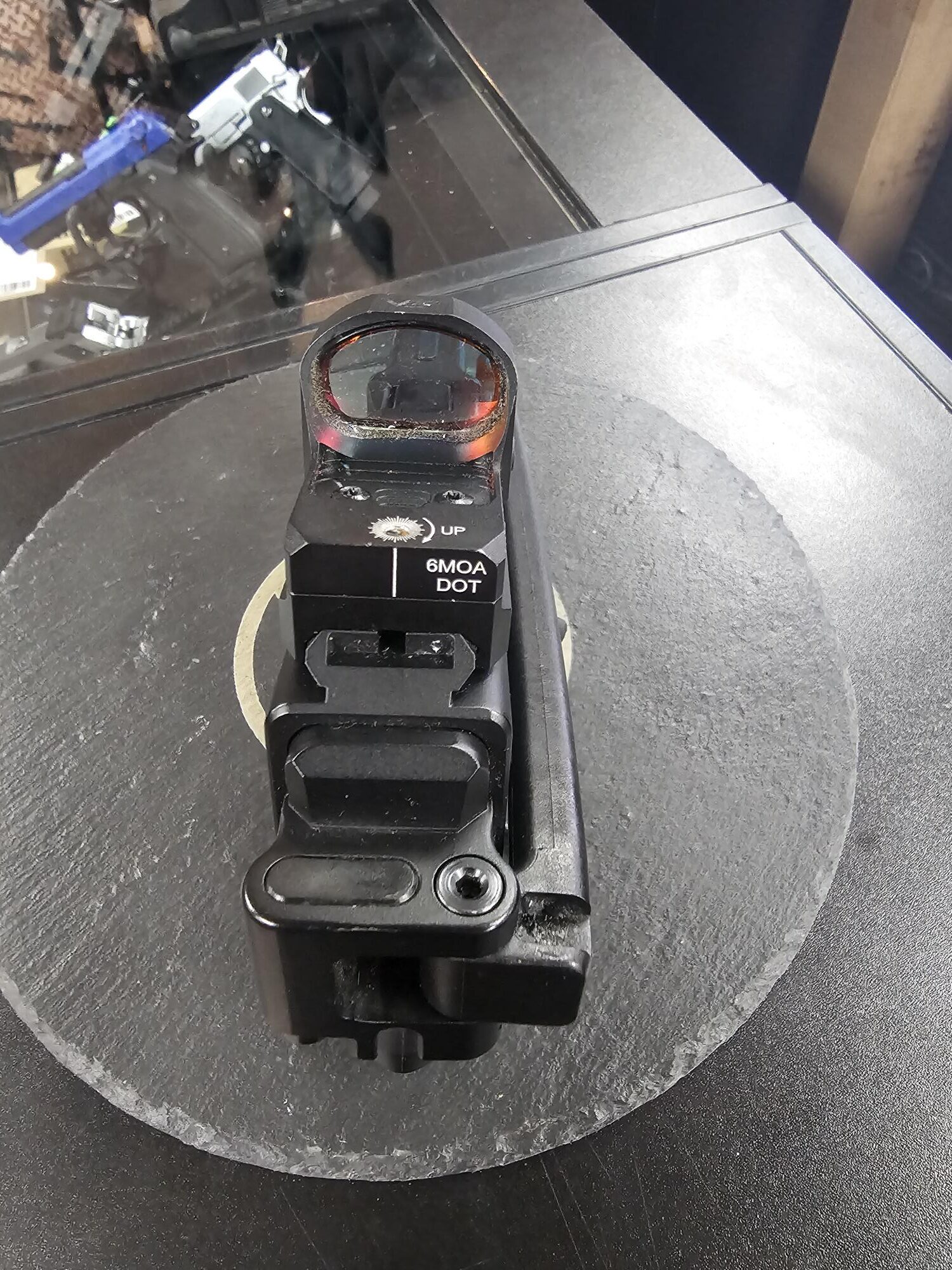 A pre-owned B&T USW airsoft pistol with a mounted red dot sight rests on a round display stand above a shop counter.
