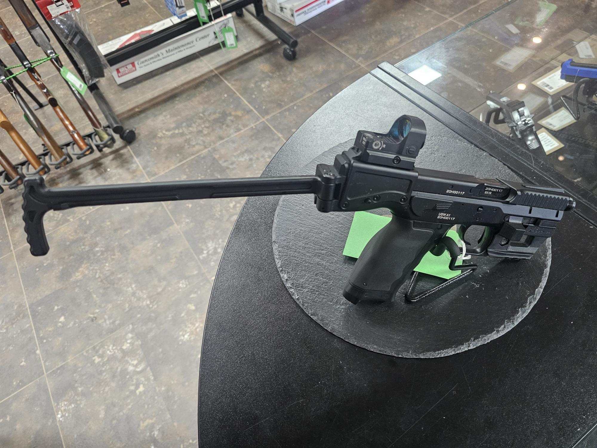Displayed on a shop counter, a pre-owned B&T USW CO2 airsoft pistol has a long folding stock and mounted red dot sight.